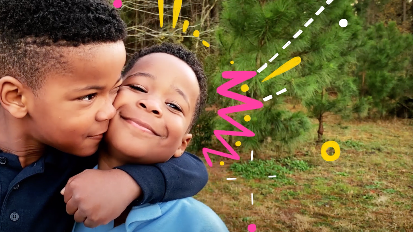 Two African-American boys embrace in front of some trees.
